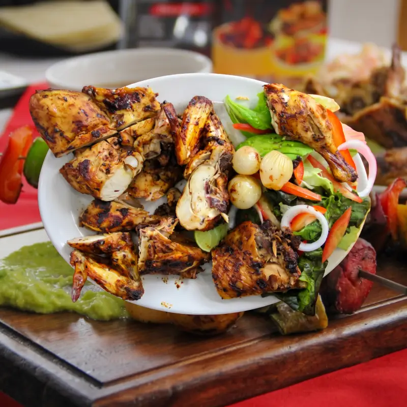 Primer plano de un plato blanco rebosante de jugosas piezas de pollo asado al carbón con textura dorada, acompañado de una ensalada fresca con lechuga, cebolla morada, pimiento rojo y cebollitas cocktail asadas, servido en el restaurante Pollo Moyo de Martínez de la Torre.