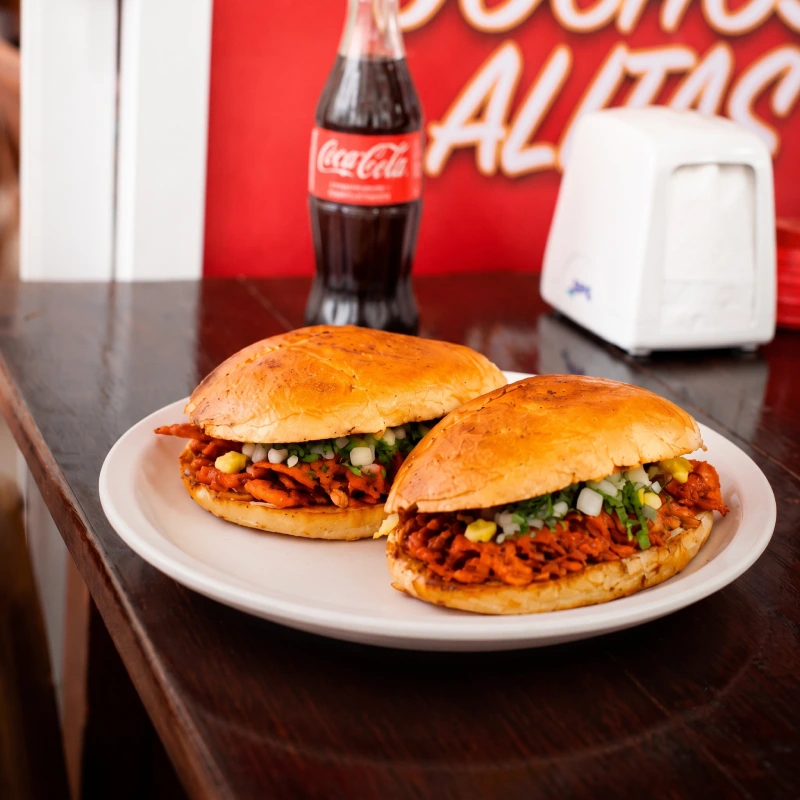 Dos tortas gigantes de carne al pastor con piña y cilantro servidas en pan doradito, junto a un refresco de cola en la taquería Taco Moyo de Martínez de la Torre.